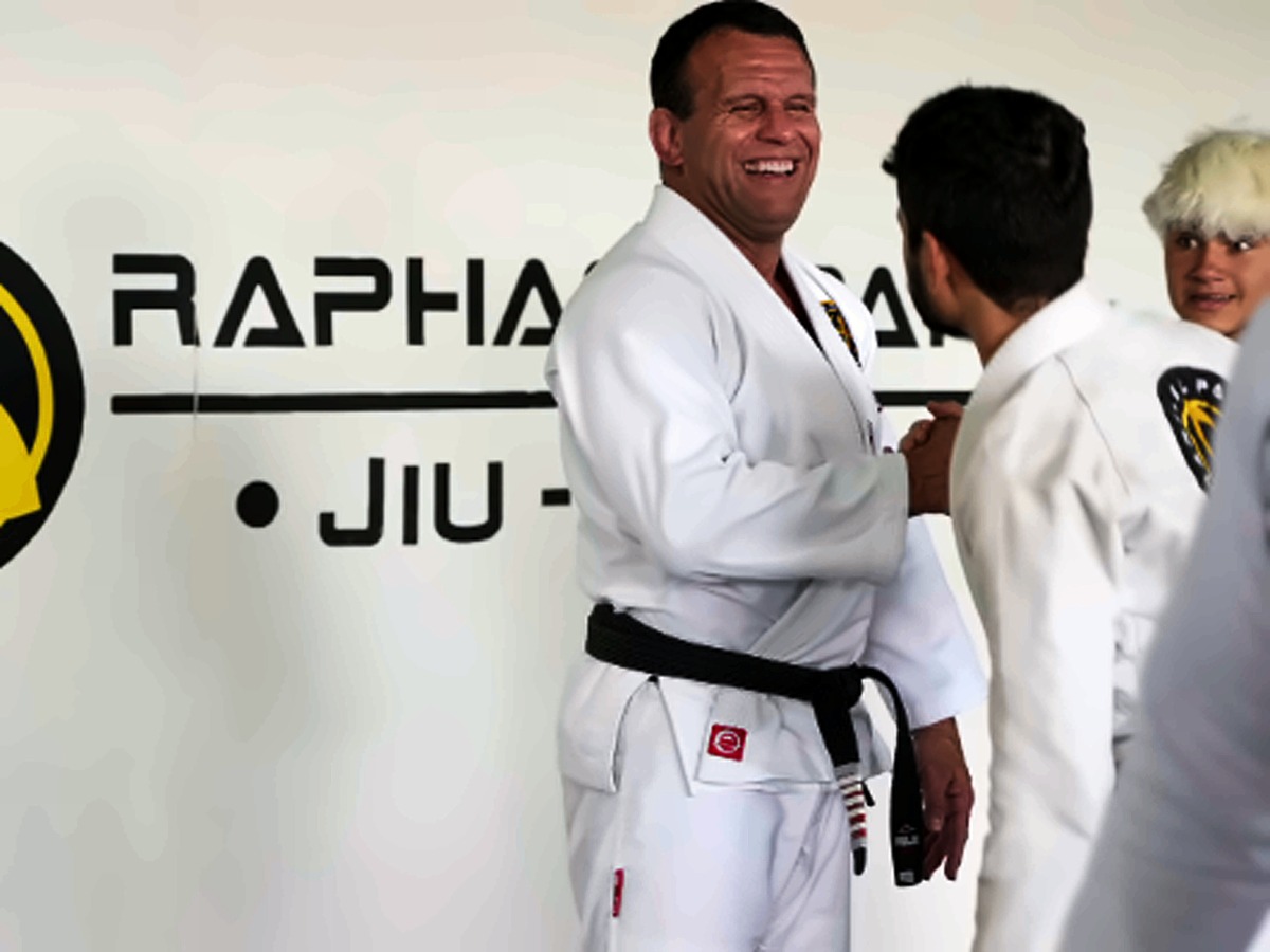 Aluno faixa preta sorrindo diante da parede com o logo do Raphael Paiva Jiu-Jitsu